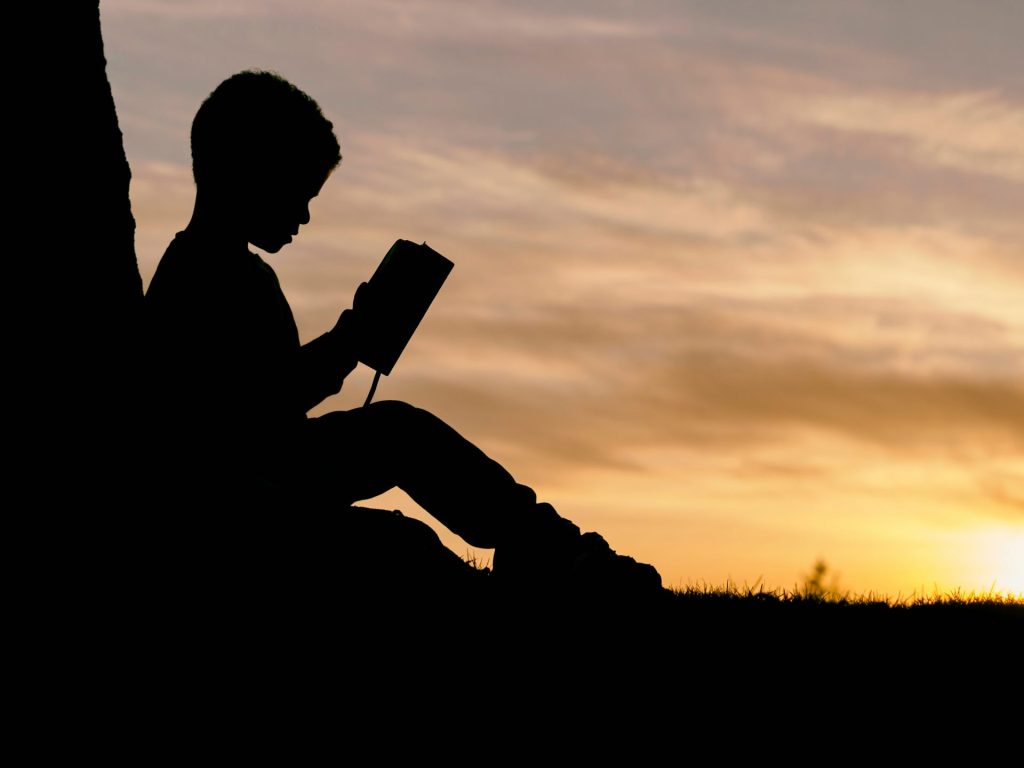 Child silhouette reading against a sunset backdrop.